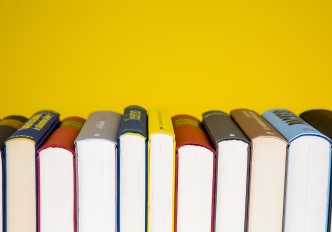 Fotografia de livros alinhados, voltados com a lombada para cima, sob fundo colorido amarelo