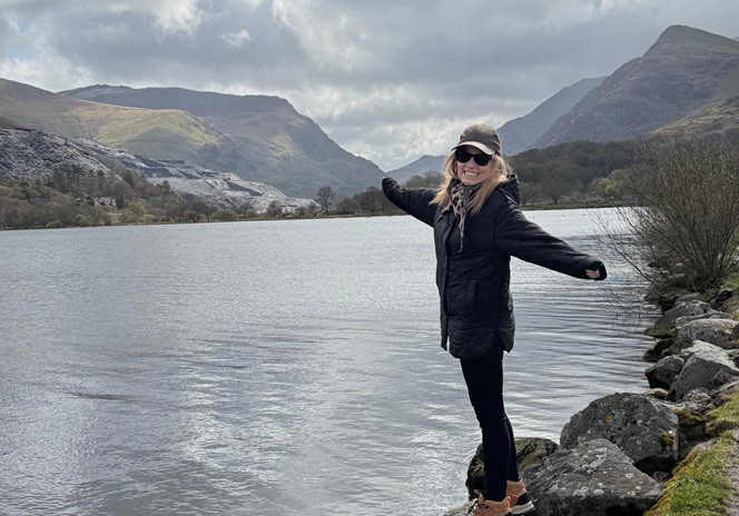 Fotografia de Maria Inês Almeida à beira de um lago com montanhas em fundo