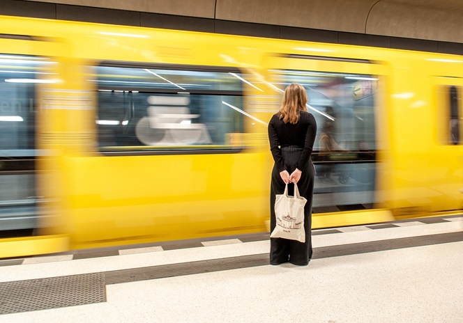 Fotografia de mulher de costas, com mochila na mão, na plataforma de comboios, com um comboio amarelo em andamento em segundo plano 